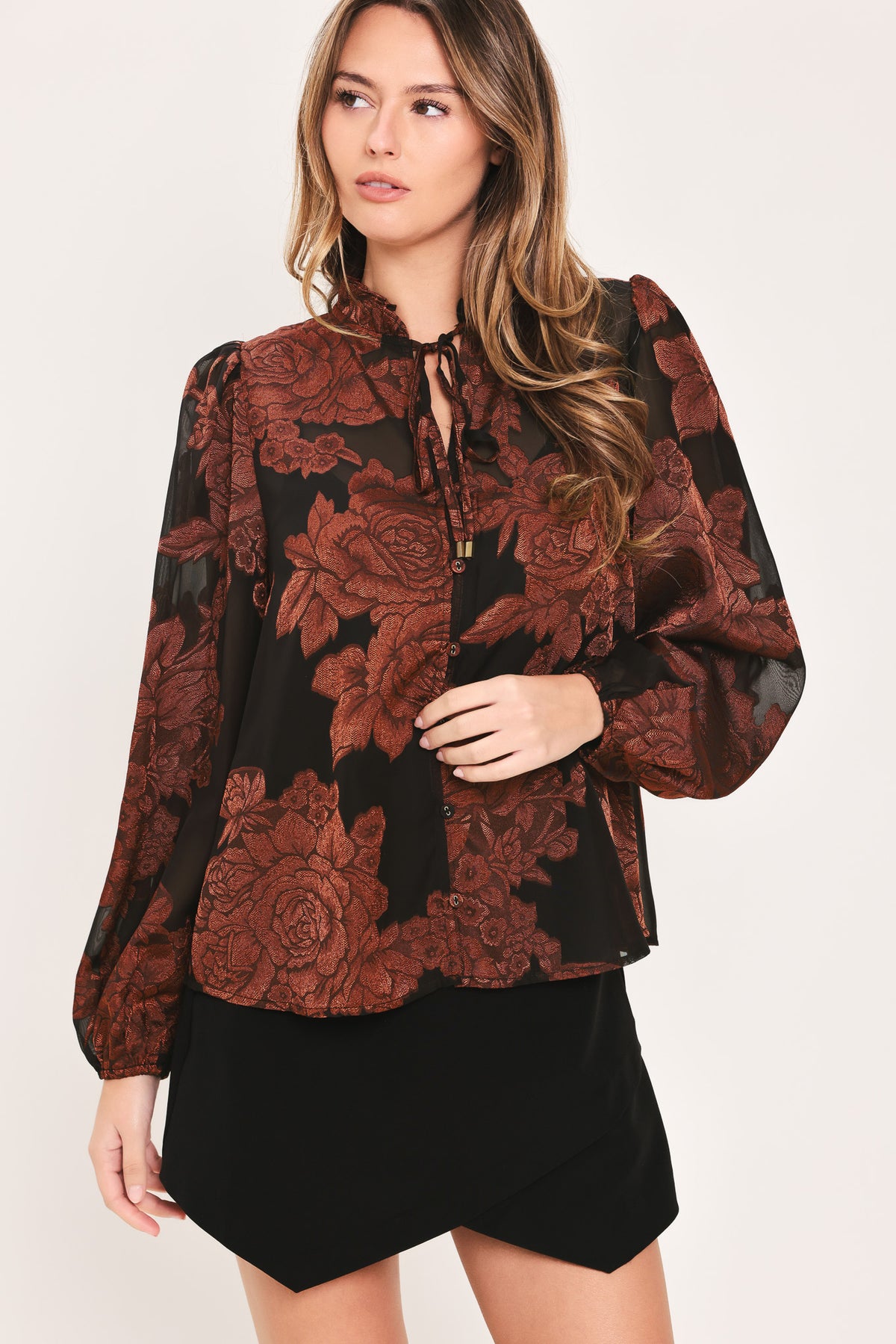 Woman wearing a black long sleeve with burgundy floral jacquard print, ruffle neck, and string tie at the center clavicle | Flying Tomato