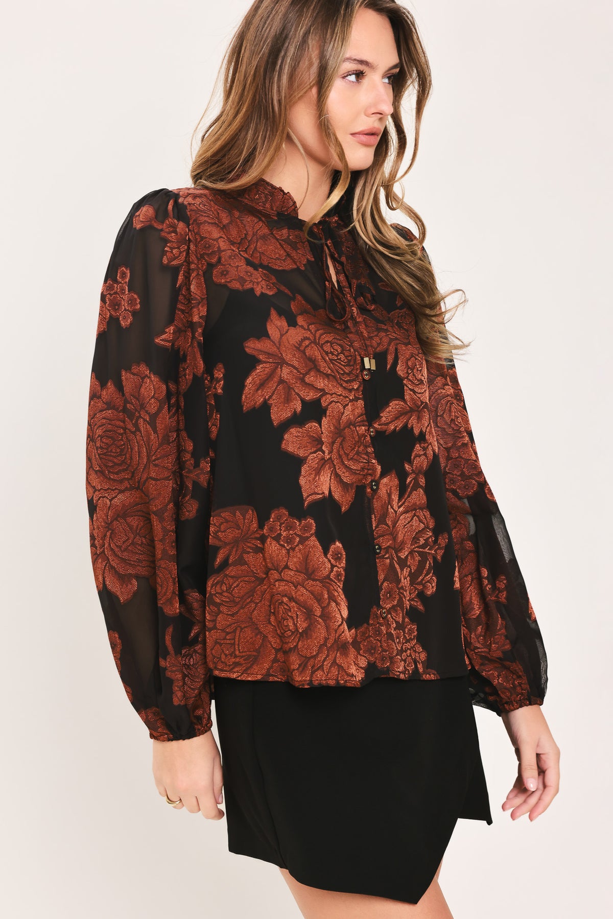 Woman wearing a black long sleeve with burgundy floral jacquard print, ruffle neck, and string tie at the center clavicle | Flying Tomato