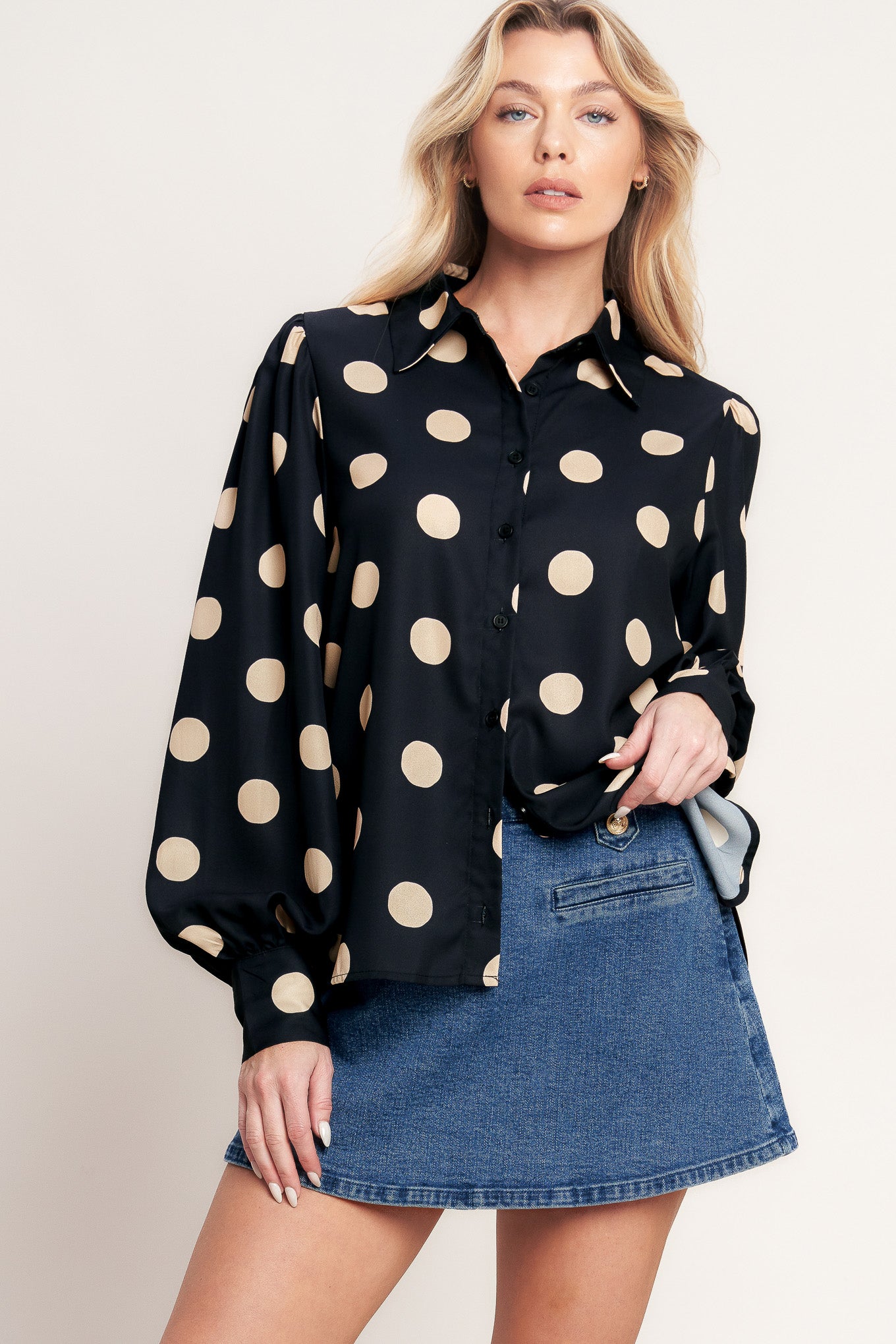 A woman modeling a black long-sleeve button-up blouse with large beige polka dots, paired with a blue denim mini skirt, against a plain light beige studio background.