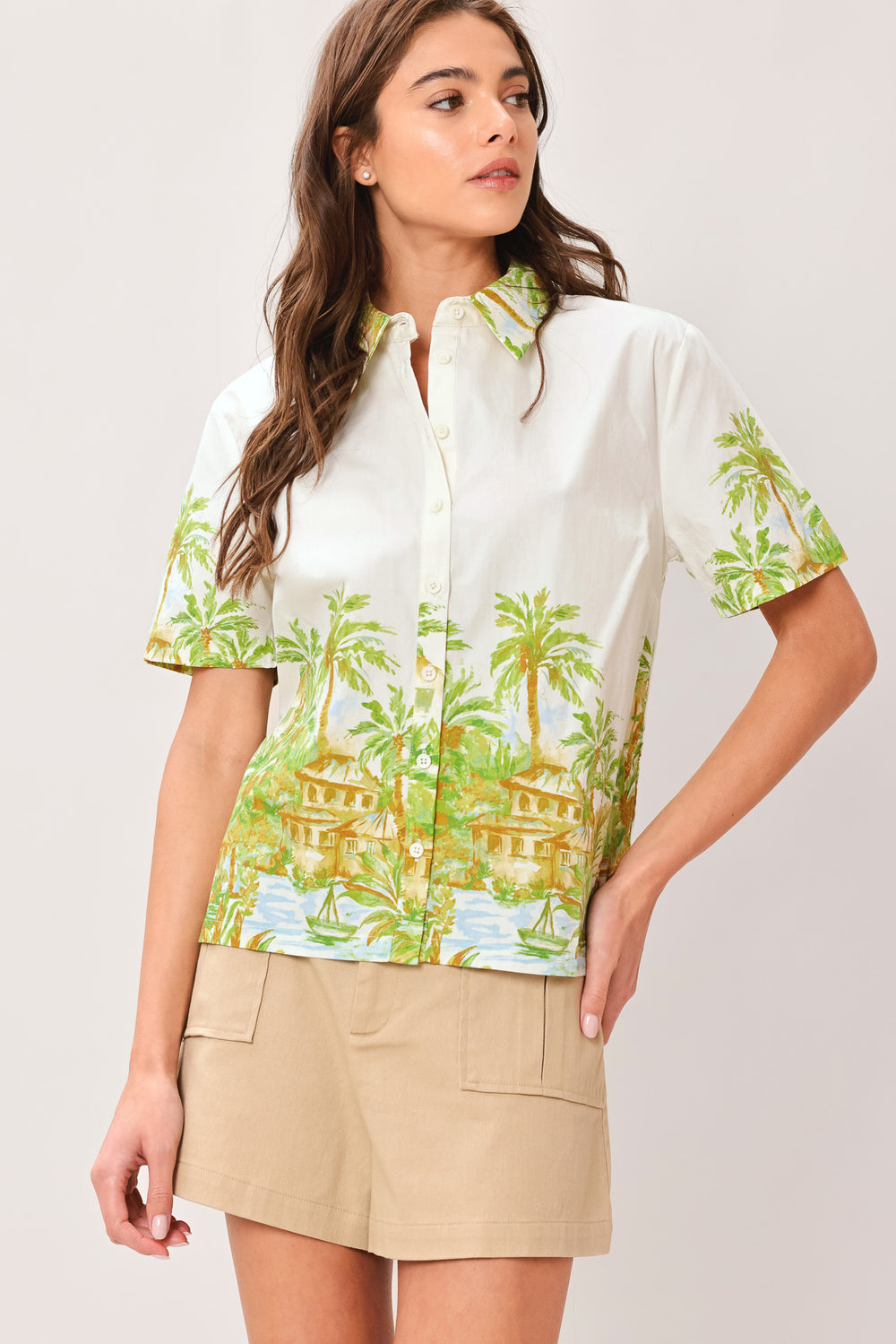 Woman wearing a white collared , short sleeve shirt with tropical print and beige shorts on a plain background | Flying Tomato