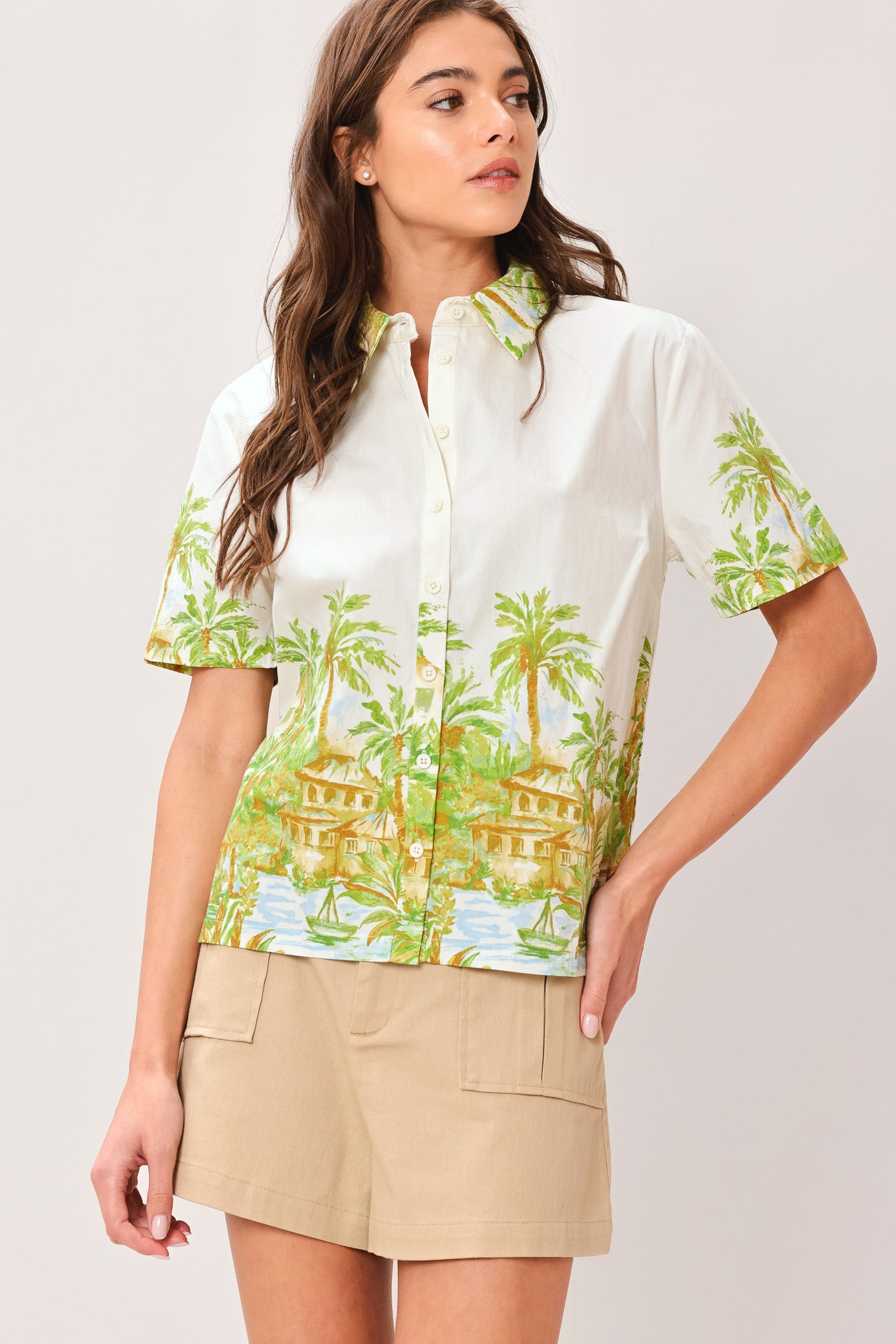 Woman wearing a white collared , short sleeve shirt with tropical print and beige shorts on a plain background | Flying Tomato