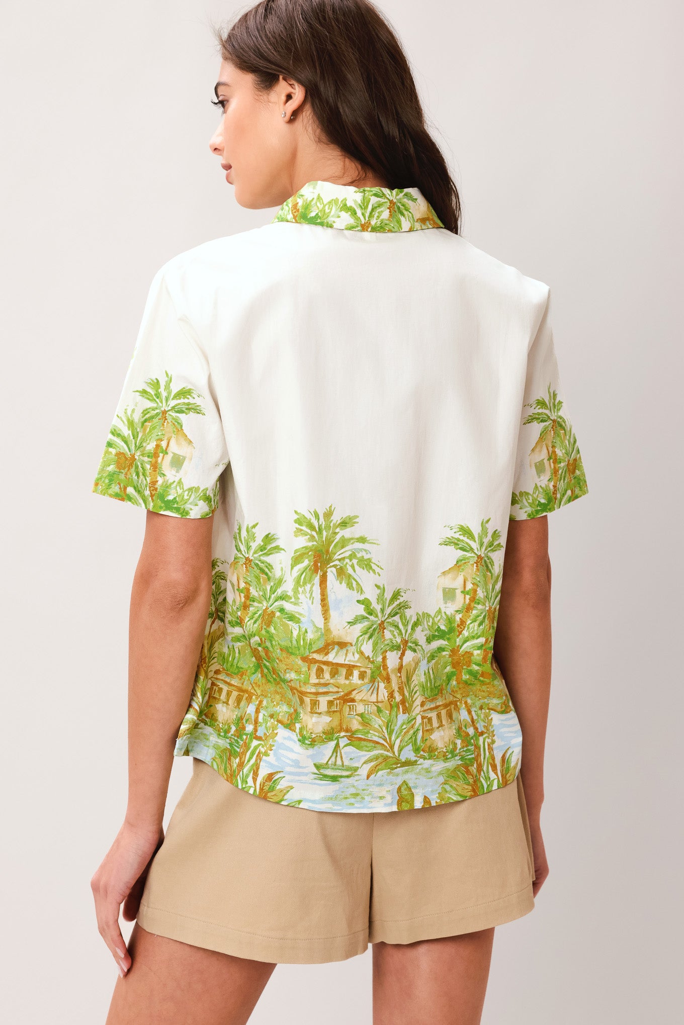 Woman wearing a white collared , short sleeve shirt with tropical print and beige shorts on a plain background | Flying Tomato
