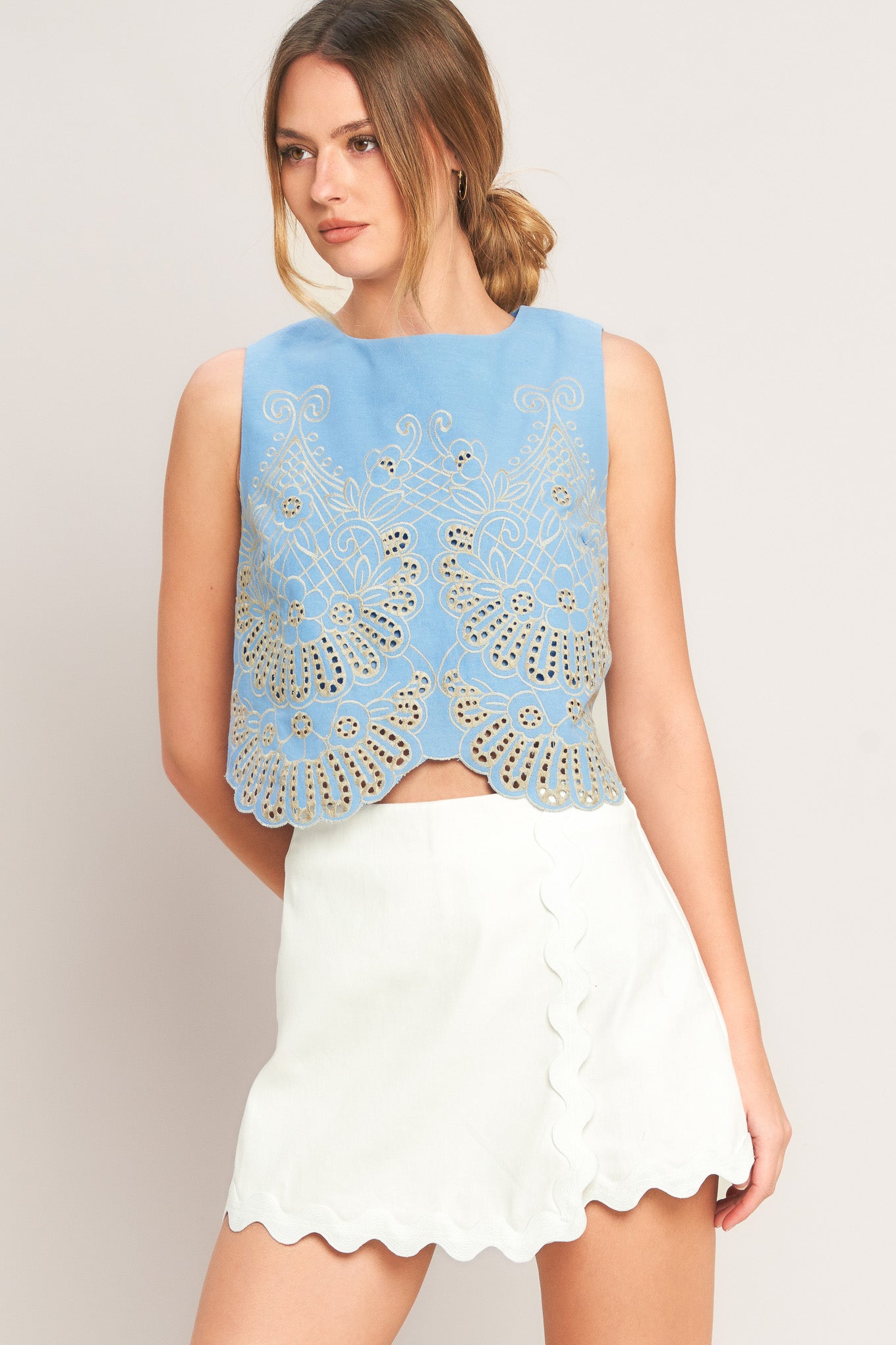 Woman wearing a light blue sleeveless top with intricate lace detailing and a white skirt on a plain background