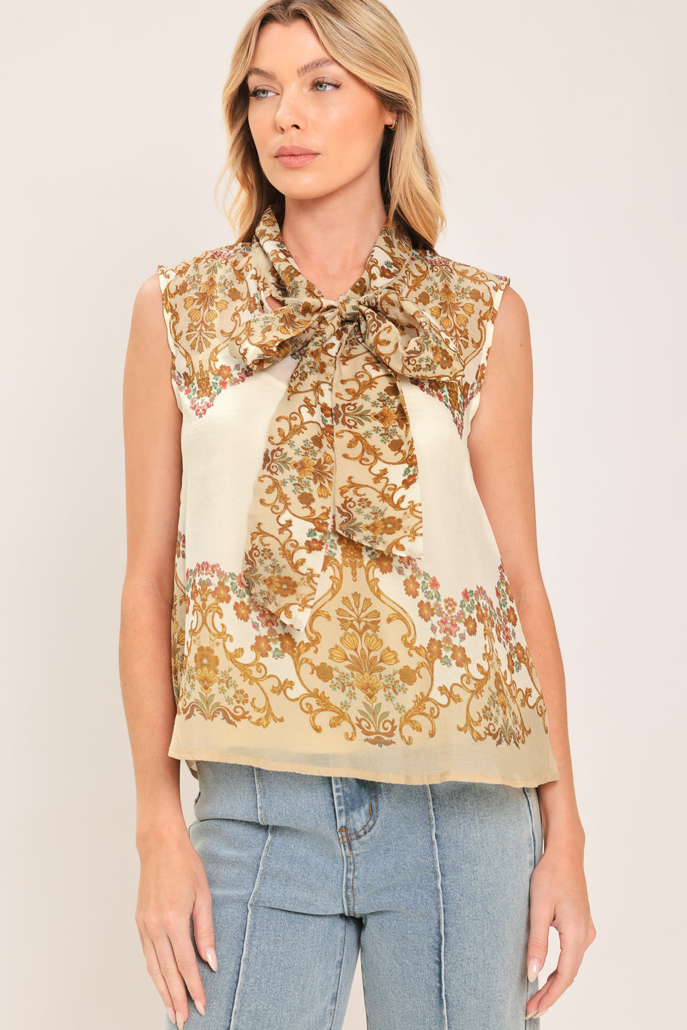 A woman wearing a sleeveless cream-colored blouse with a brown and gold damask pattern and a large bow tie-neck | Flying Tomato