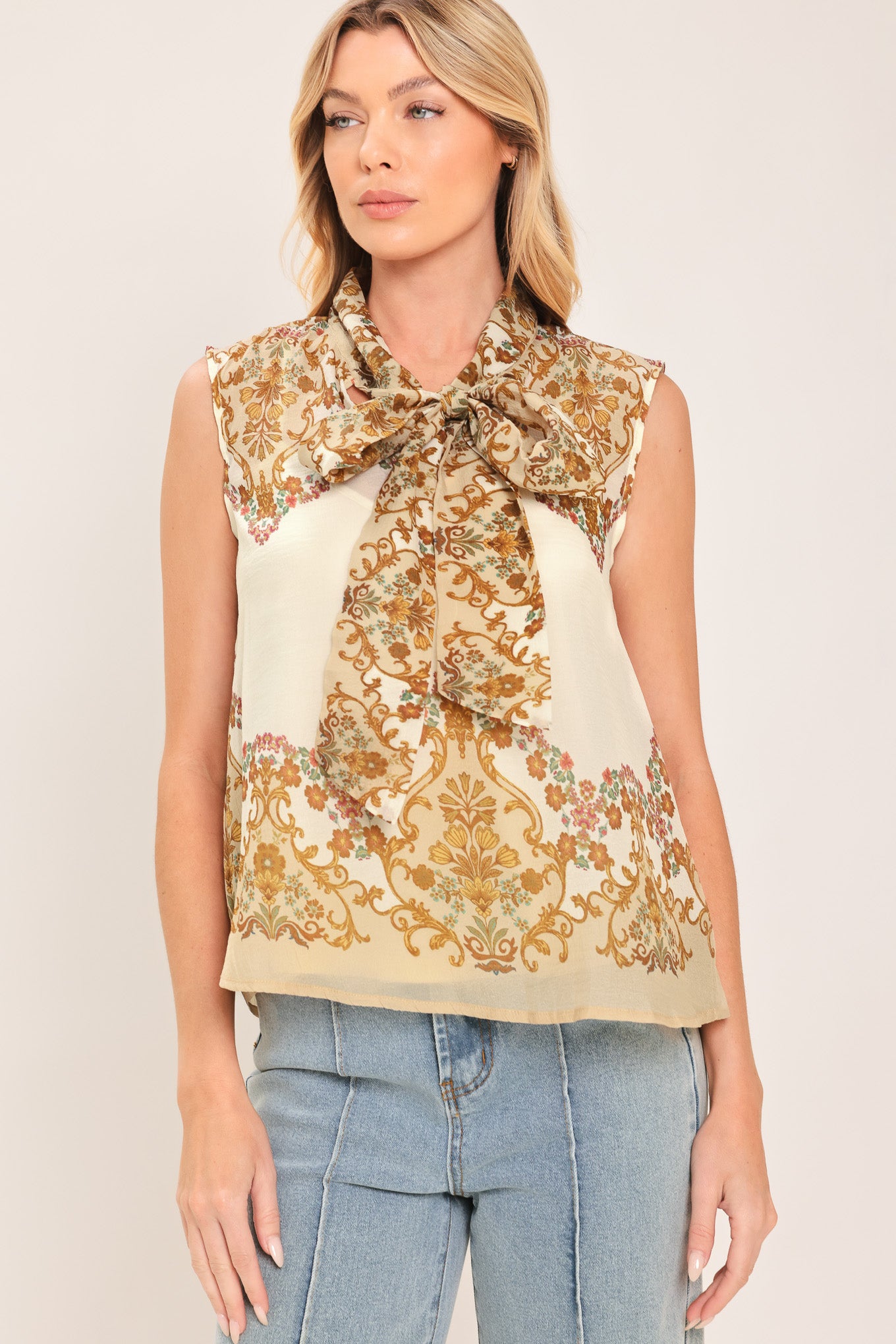 A woman wearing a sleeveless cream-colored blouse with a brown and gold damask pattern and a large bow tie-neck | Flying Tomato