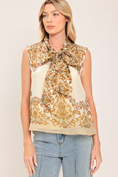 A woman wearing a sleeveless cream-colored blouse with a brown and gold damask pattern and a large bow tie-neck | Flying Tomato