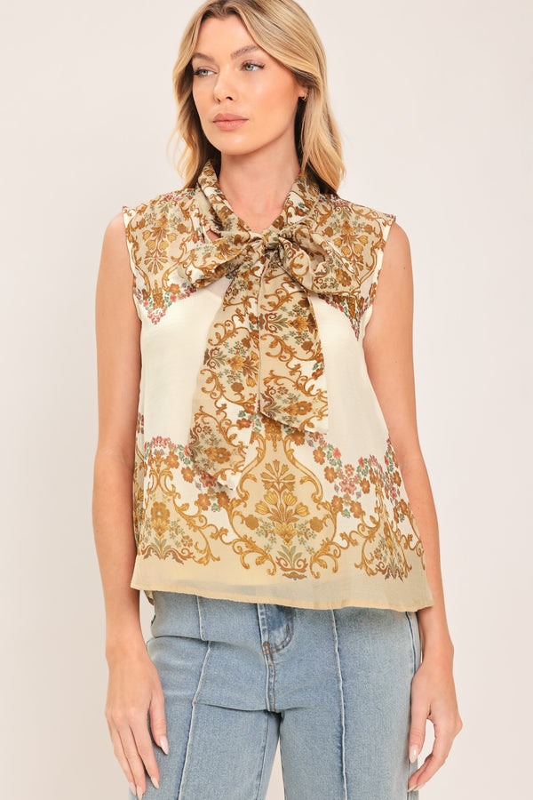 A woman wearing a sleeveless cream-colored blouse with a brown and gold damask pattern and a large bow tie-neck | Flying Tomato