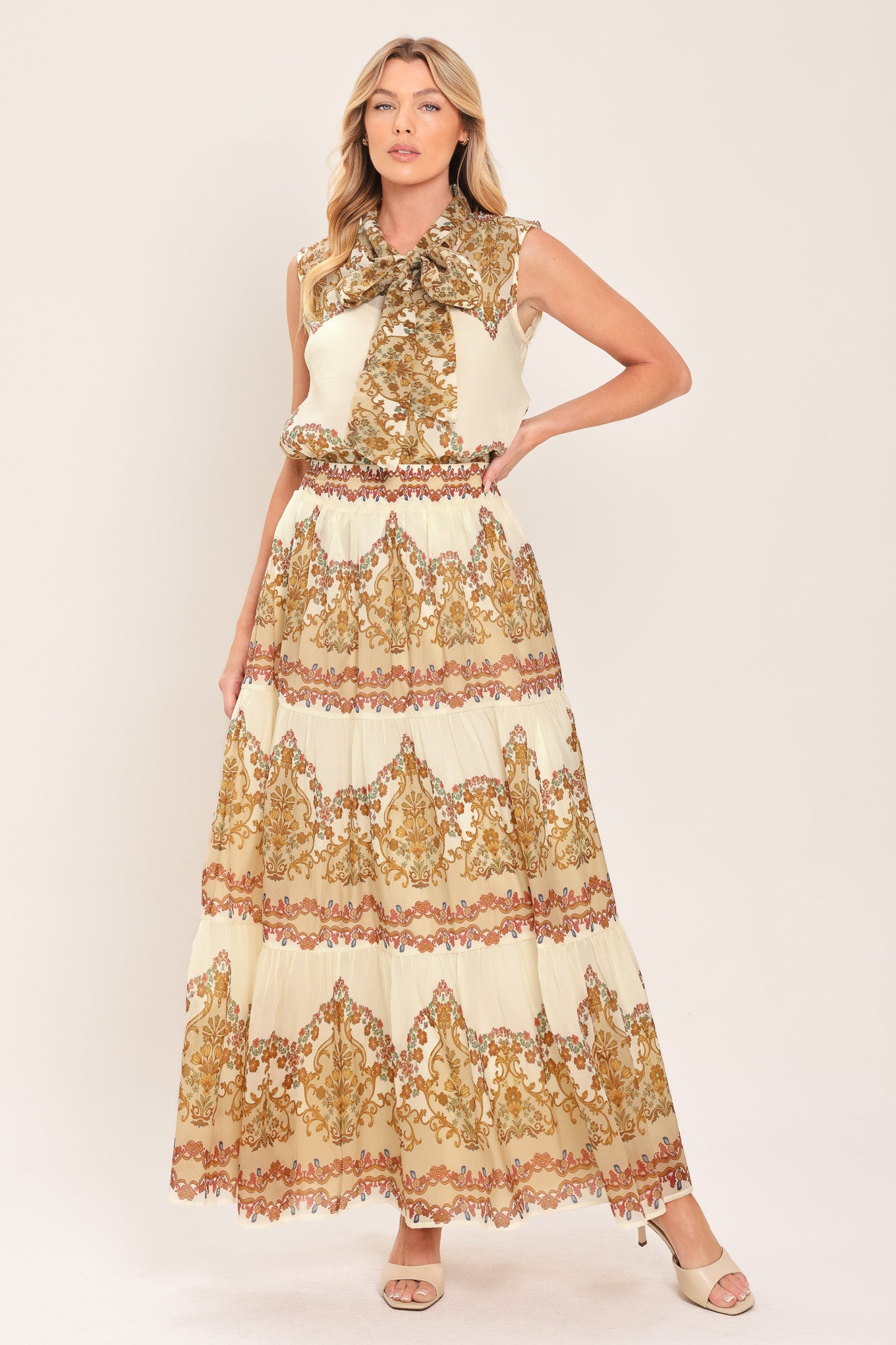 A woman wearing a sleeveless cream-colored blouse with a brown and gold damask pattern and a large bow tie-neck | Flying Tomato