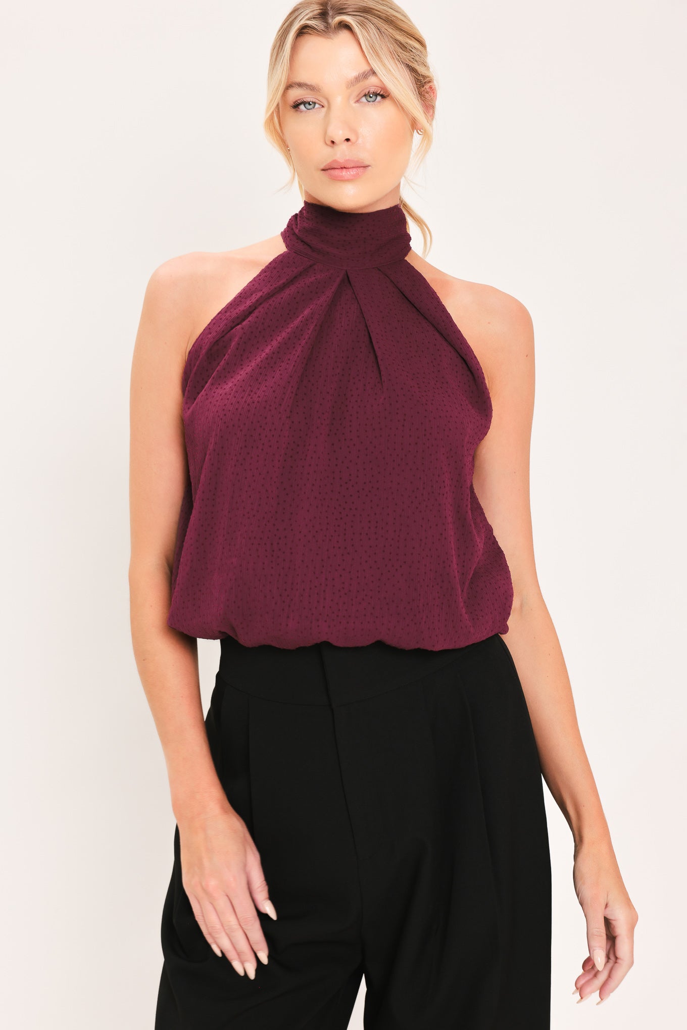 A woman modeling a sleeveless halter-style knit top in deep burgundy with a gathered high neck and scalloped hem | Flying Tomato