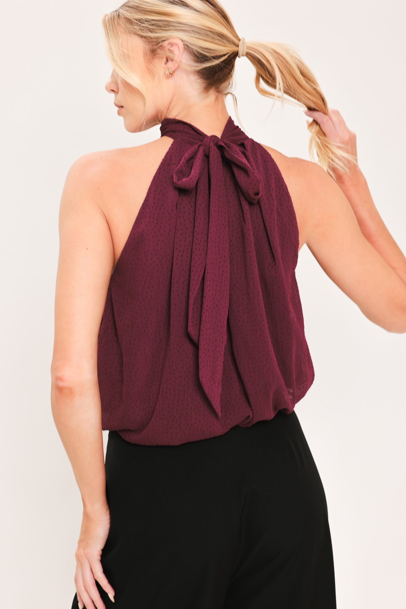 A woman modeling a sleeveless halter-style knit top in deep burgundy with a gathered high neck and scalloped hem | Flying Tomato