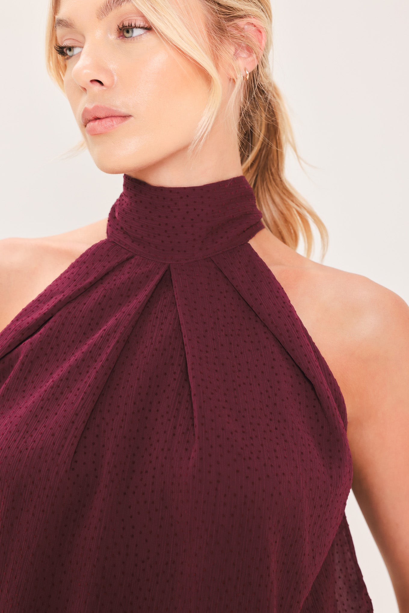 A woman modeling a sleeveless halter-style knit top in deep burgundy with a gathered high neck and scalloped hem | Flying Tomato
