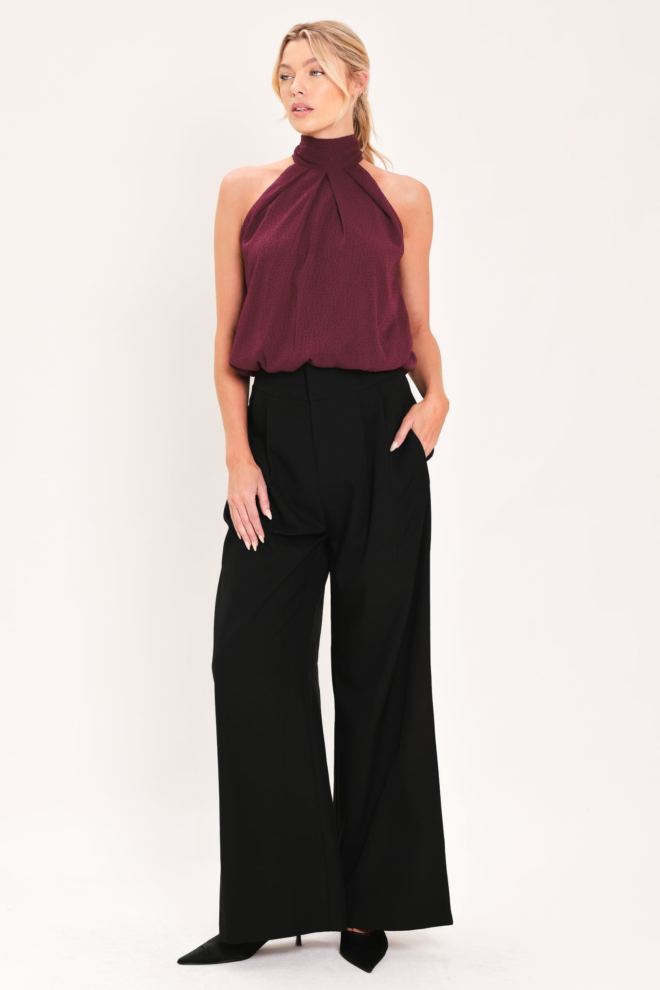 A woman modeling a sleeveless halter-style knit top in deep burgundy with a gathered high neck and scalloped hem | Flying Tomato