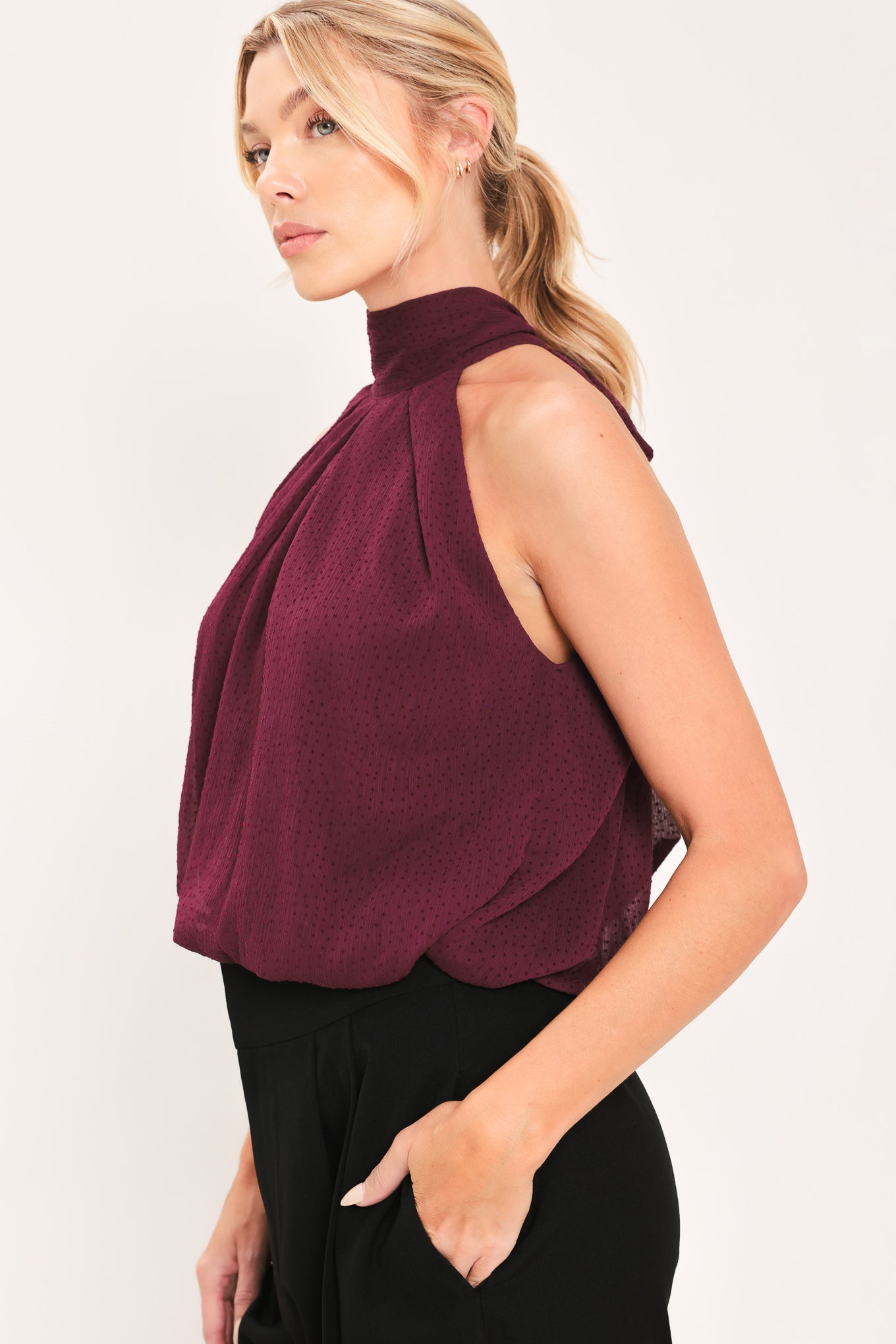 A woman modeling a sleeveless halter-style knit top in deep burgundy with a gathered high neck and scalloped hem | Flying Tomato