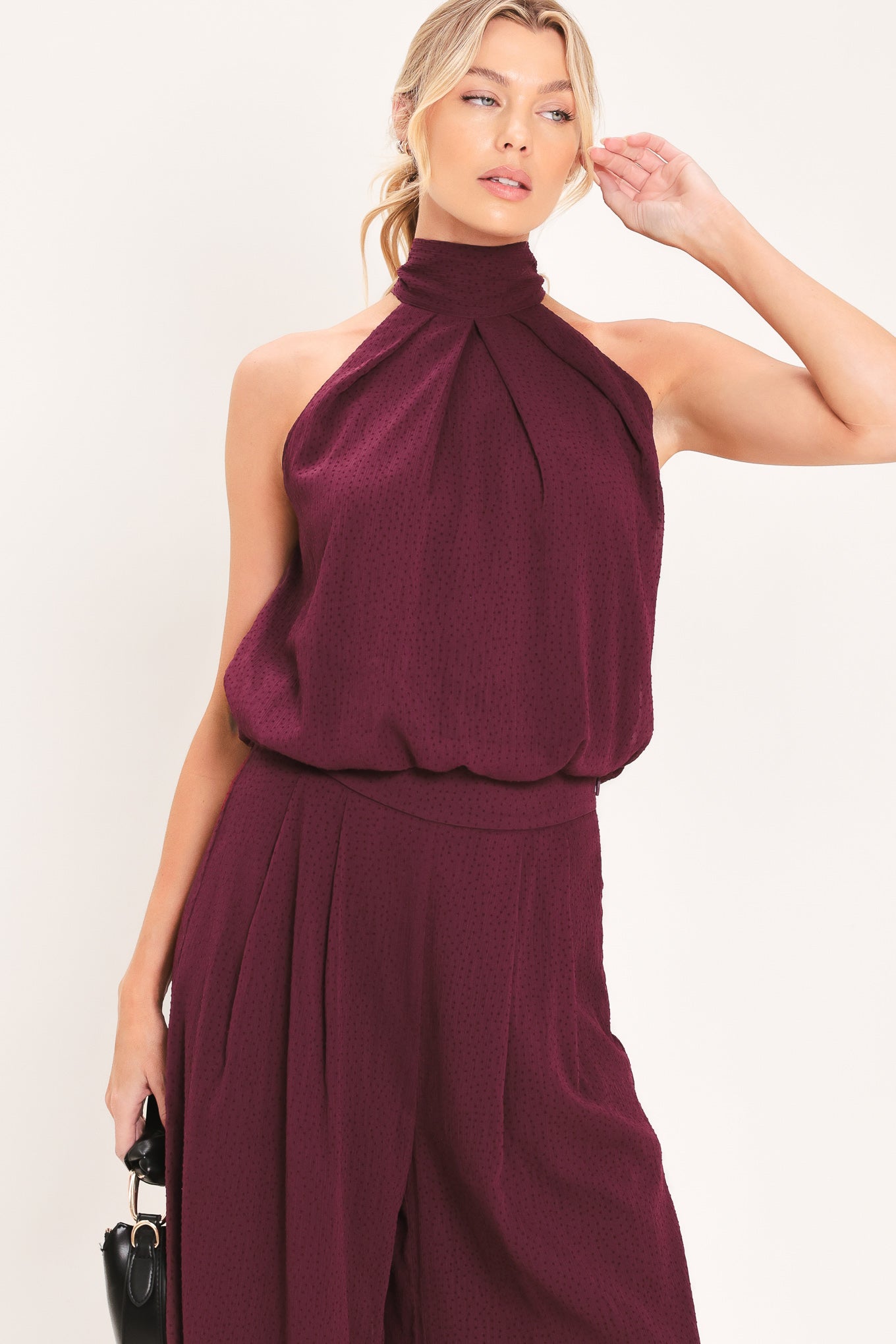 A woman modeling a sleeveless halter-style knit top in deep burgundy with a gathered high neck and scalloped hem | Flying Tomato