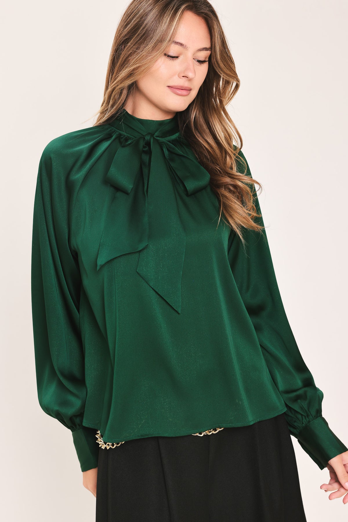 A woman wearing a dark hunter green satin blouse with a high neck, large bow at the collar, and gathered long sleeves with gold button accents | Flying Tomato