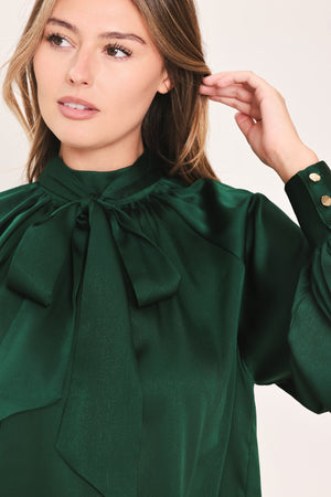 A woman wearing a dark hunter green satin blouse with a high neck, large bow at the collar, and gathered long sleeves with gold button accents | Flying Tomato