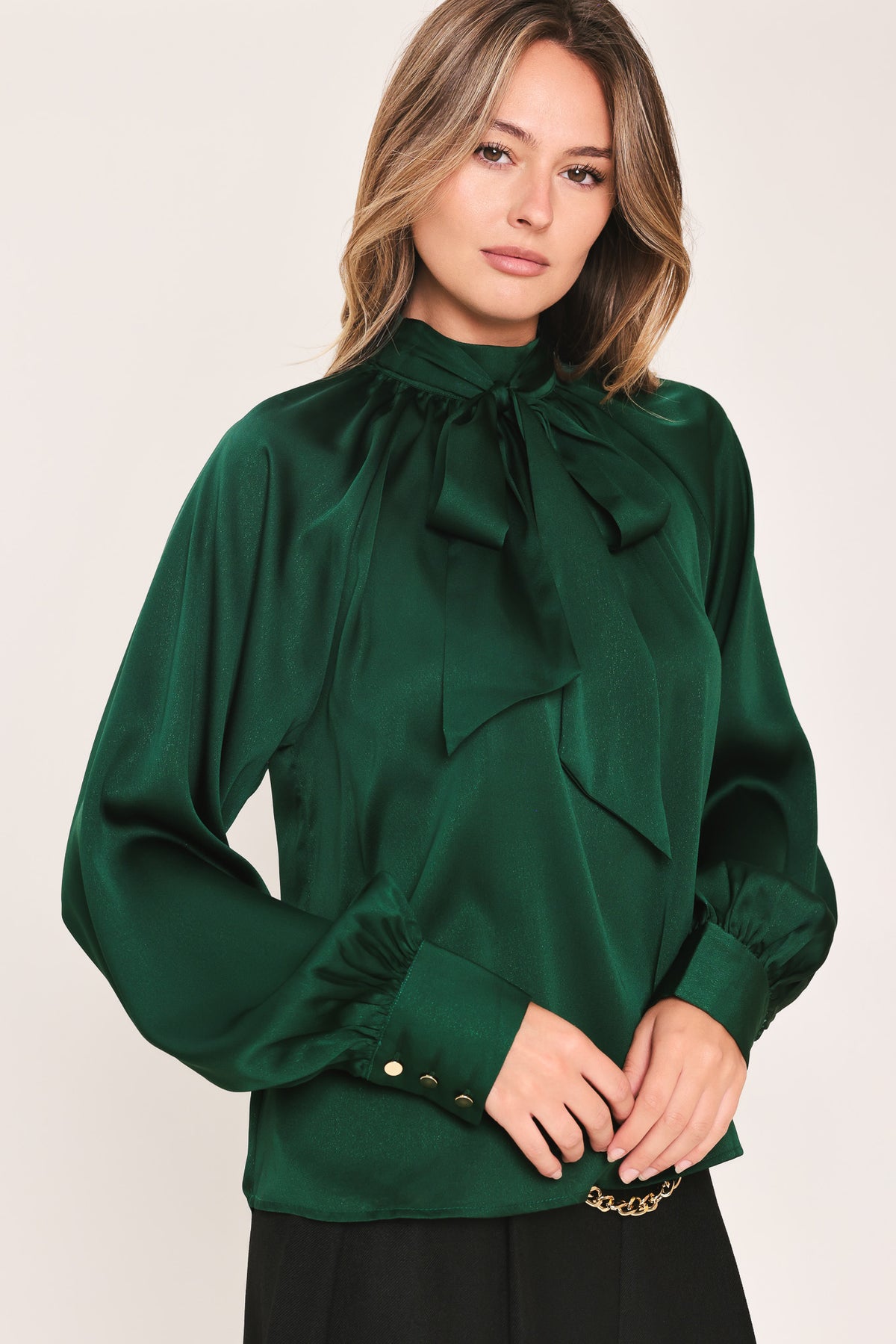 A woman wearing a dark hunter green satin blouse with a high neck, large bow at the collar, and gathered long sleeves with gold button accents | Flying Tomato