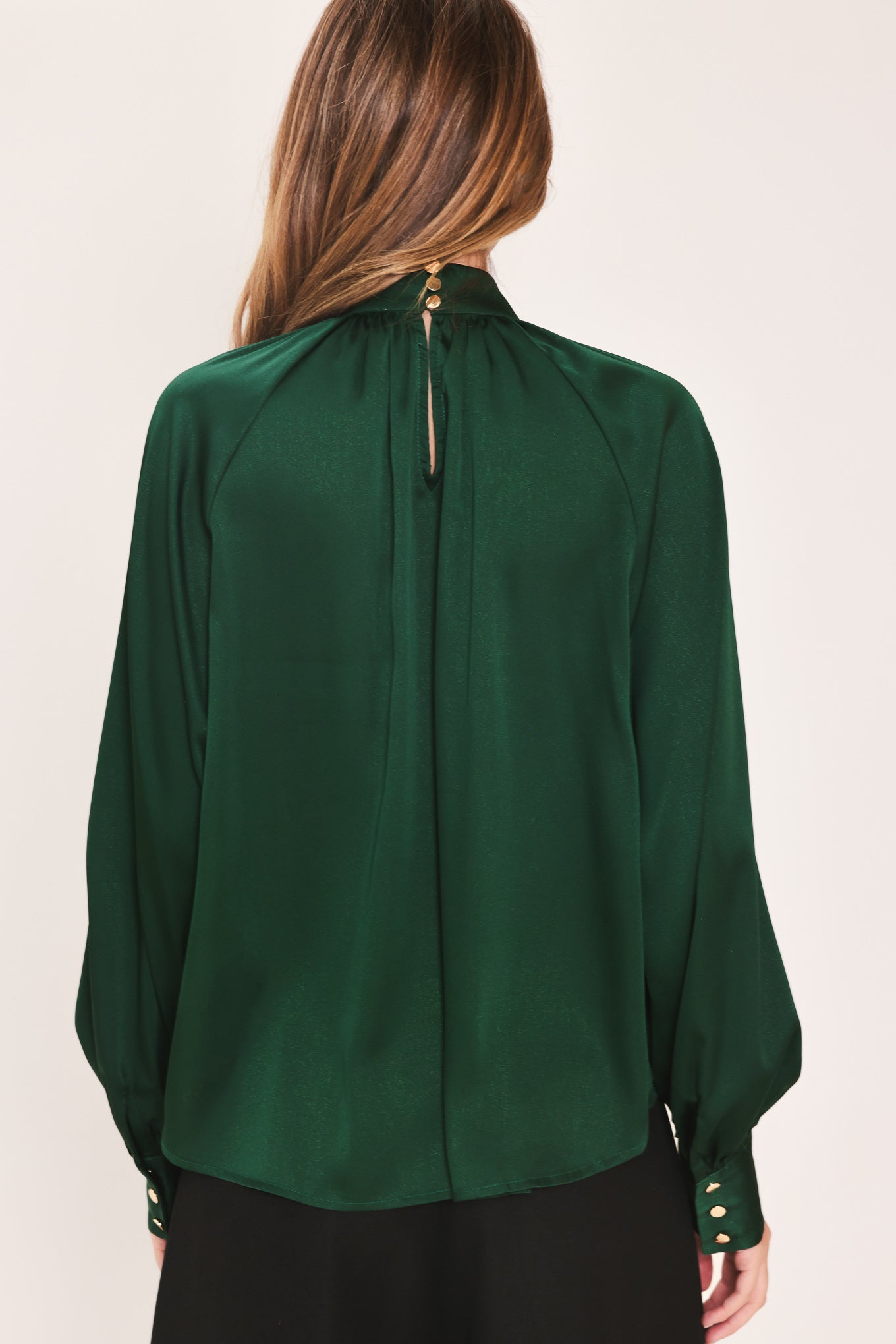 A woman wearing a dark hunter green satin blouse with a high neck, large bow at the collar, and gathered long sleeves with gold button accents | Flying Tomato