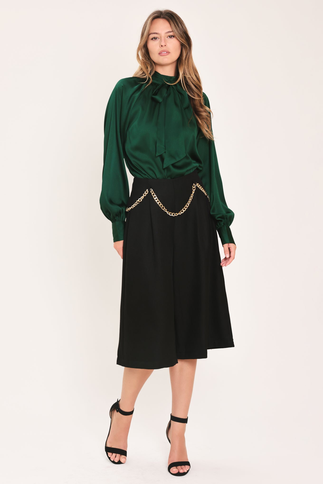 A woman wearing a dark hunter green satin blouse with a high neck, large bow at the collar, and gathered long sleeves with gold button accents | Flying Tomato
