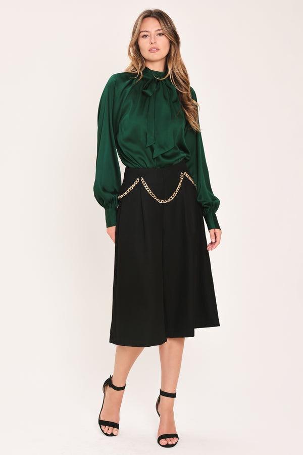 A woman wearing a dark hunter green satin blouse with a high neck, large bow at the collar, and gathered long sleeves with gold button accents | Flying Tomato