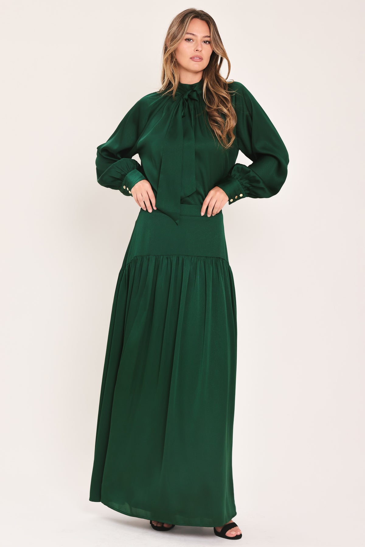A woman wearing a dark hunter green satin blouse with a high neck, large bow at the collar, and gathered long sleeves with gold button accents | Flying Tomato