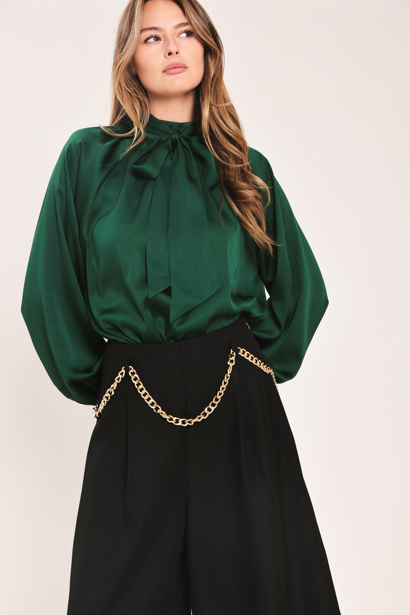 A woman wearing a dark hunter green satin blouse with a high neck, large bow at the collar, and gathered long sleeves with gold button accents | Flying Tomato