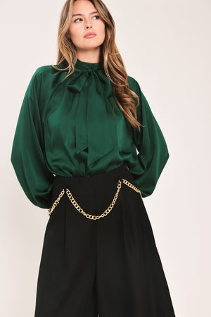 A woman wearing a dark hunter green satin blouse with a high neck, large bow at the collar, and gathered long sleeves with gold button accents | Flying Tomato