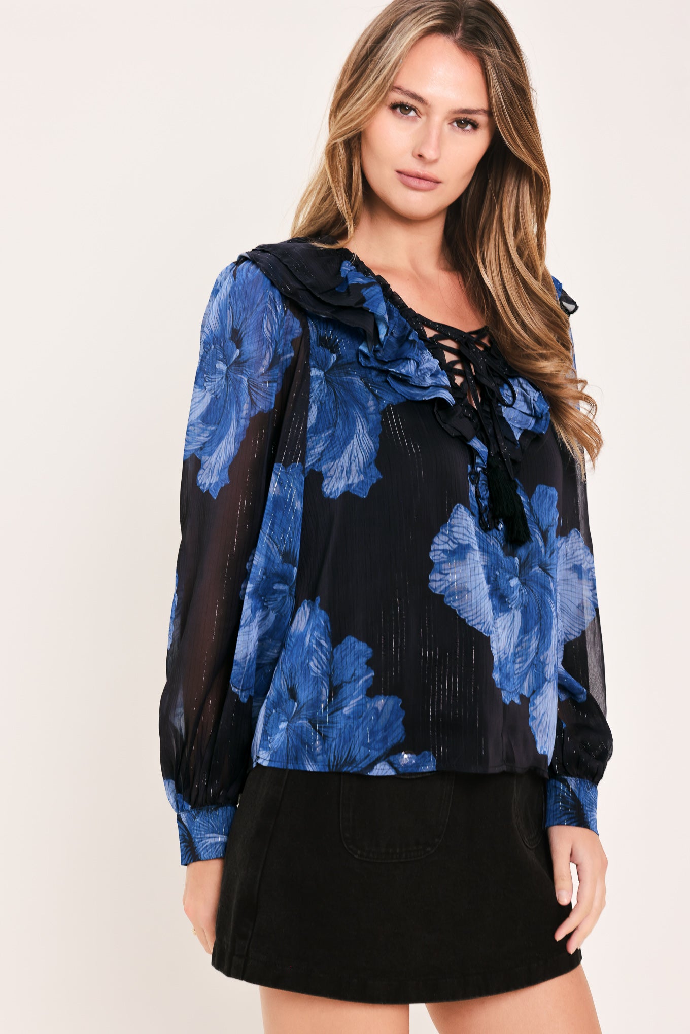 A woman wearing a sheer black blouse with a blue floral print, ruffled collar, and lace-up neckline | Flying Tomato