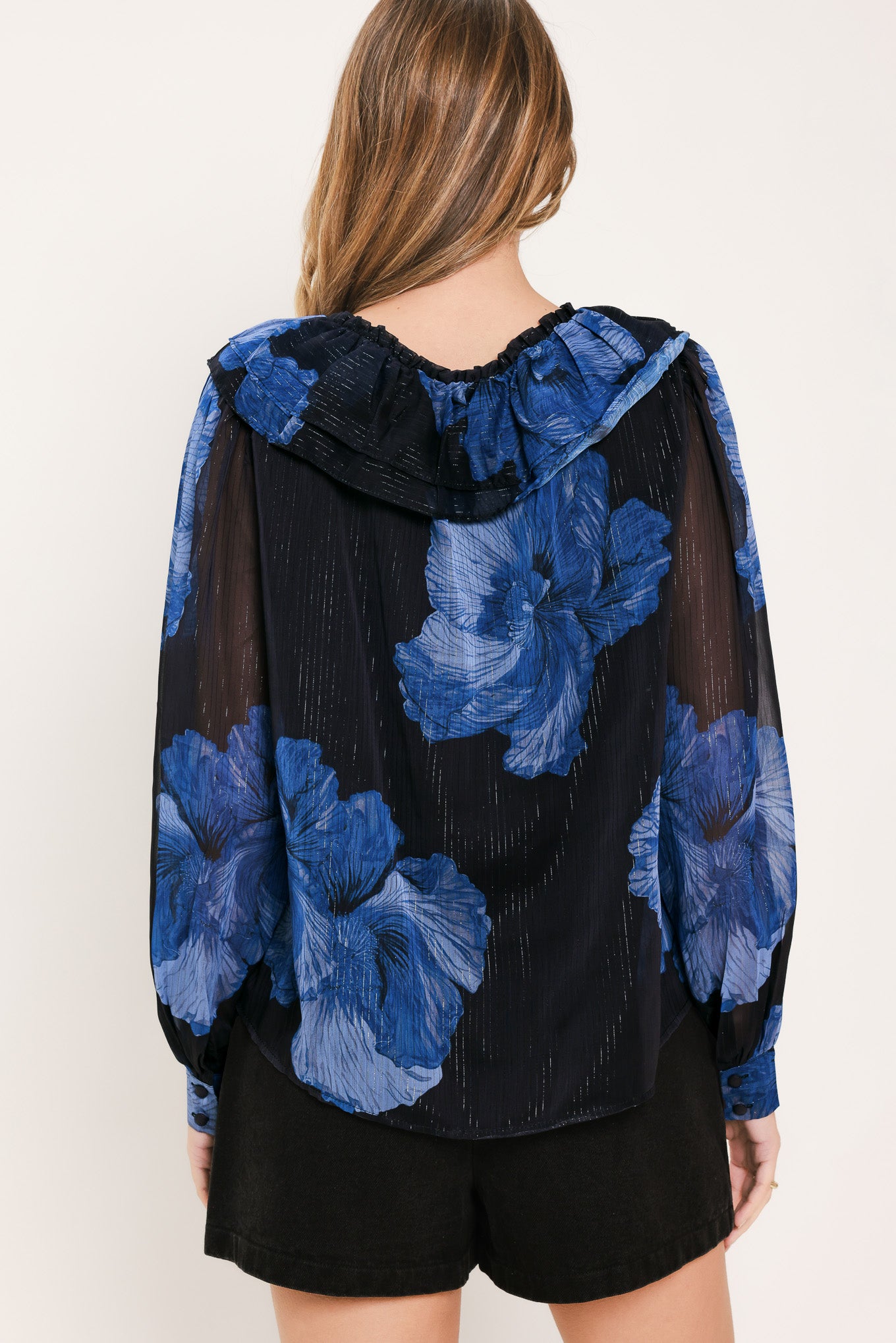 A woman wearing a sheer black blouse with a blue floral print, ruffled collar, and lace-up neckline | Flying Tomato