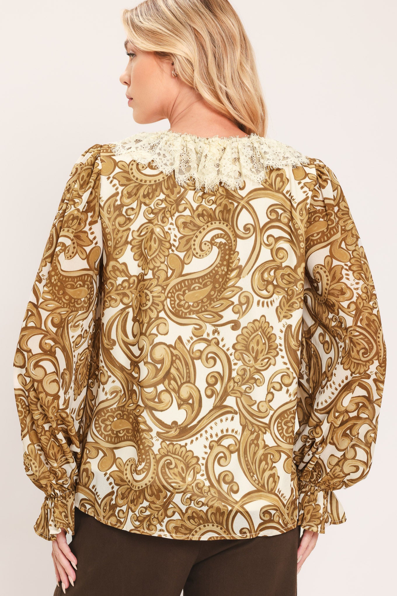A woman wearing a long-sleeve blouse with a gold and beige baroque-style pattern, a ruffled lace-up neckline, and ruffled cuffs | Flying Tomato