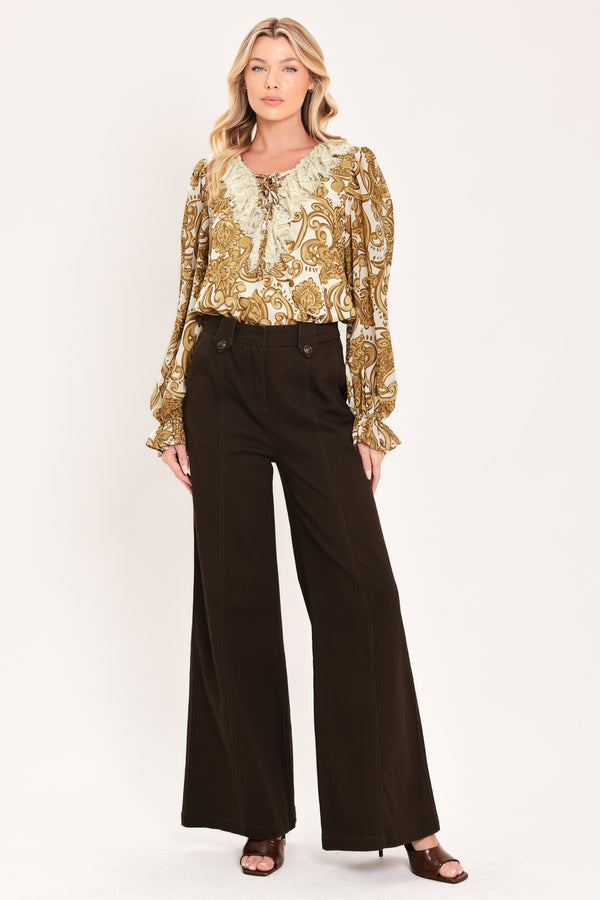 A woman wearing a long-sleeve blouse with a gold and beige baroque-style pattern, a ruffled lace-up neckline, and ruffled cuffs | Flying Tomato
