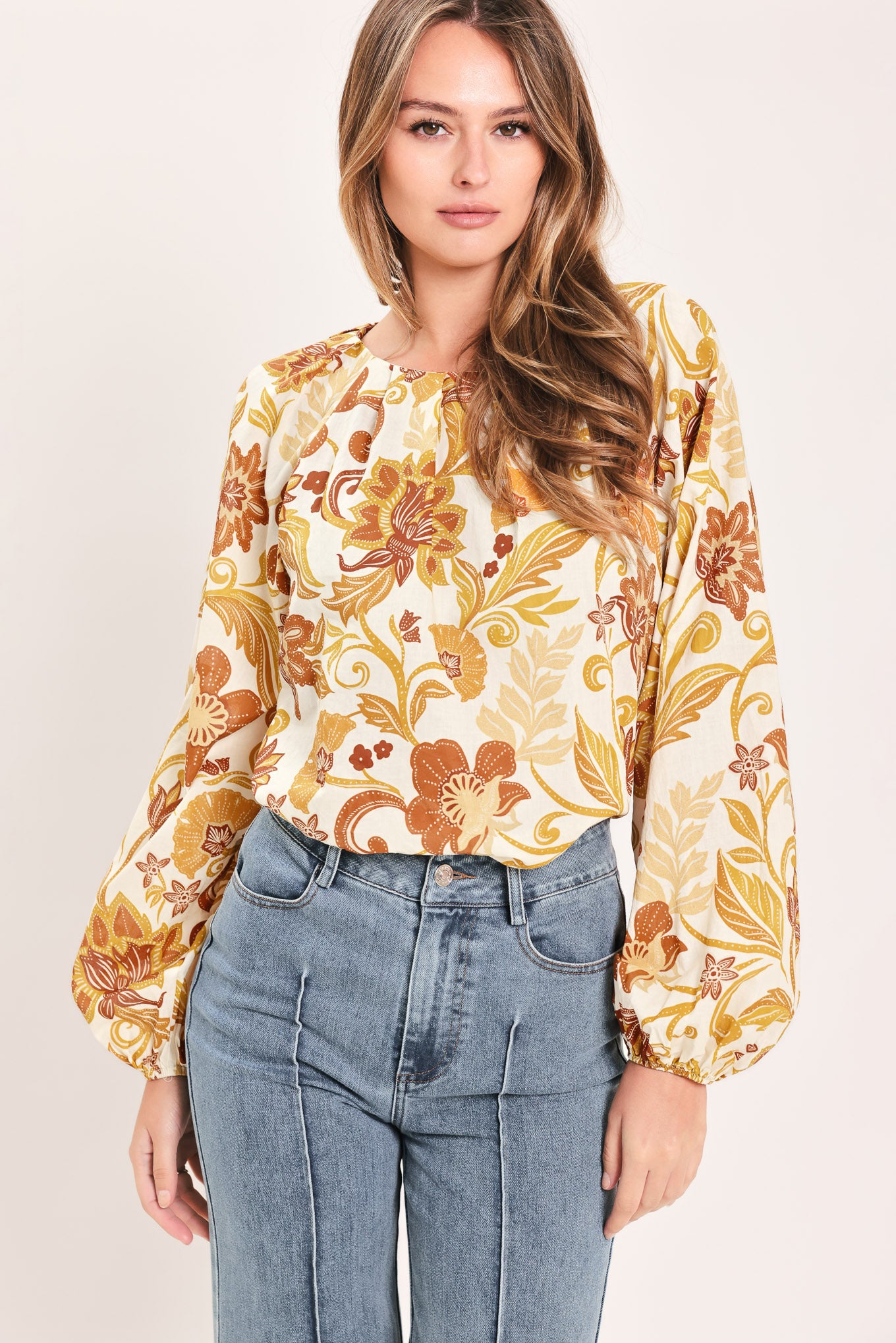 A woman wearing a long-sleeve blouse with a cream base and a brown and gold floral pattern | Flying Tomato