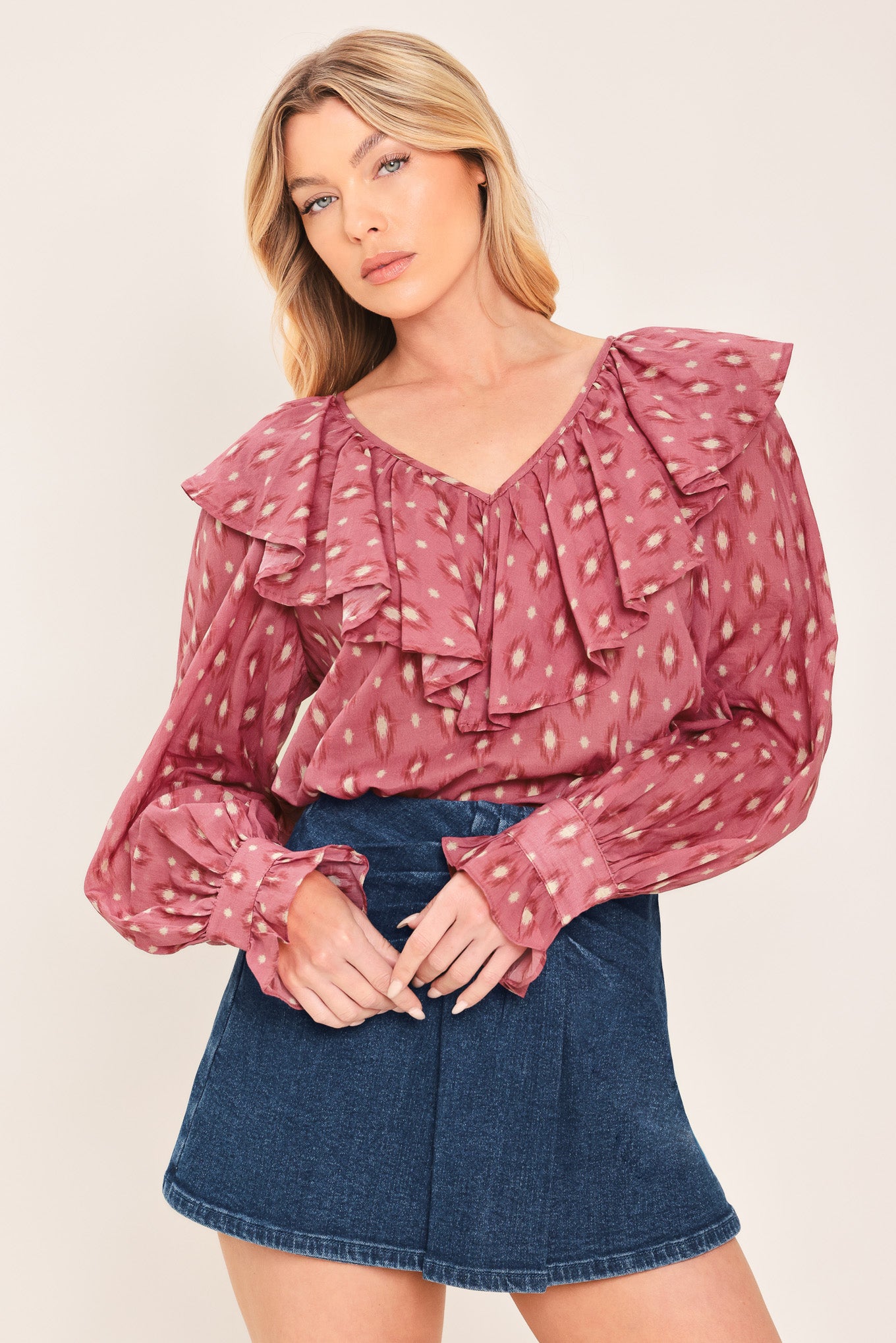 A woman wearing a dusty rose pink long-sleeve blouse with a white and beige ikat pattern, featuring layered ruffles around the V-neck and cuffs | Flying Tomato