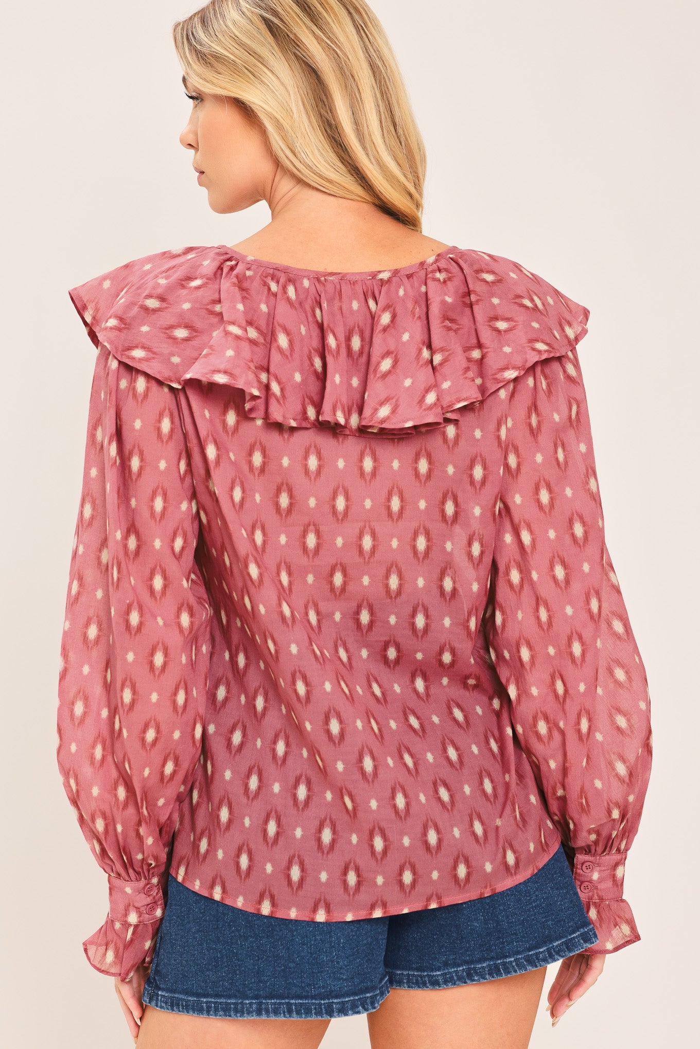 A woman wearing a dusty rose pink long-sleeve blouse with a white and beige ikat pattern, featuring layered ruffles around the V-neck and cuffs | Flying Tomato