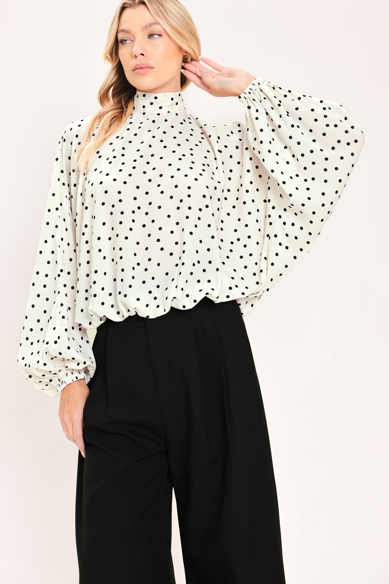A woman wearing a white long-sleeve blouse with a black polka dot pattern and a high mock neck | Flying Tomato