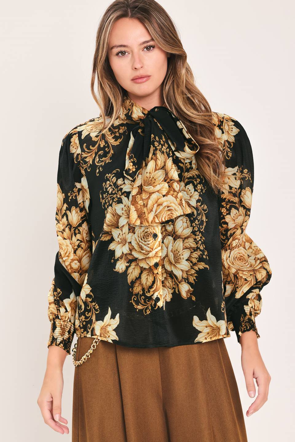 A woman wearing a black long-sleeve blouse with a large beige and gold floral print, featuring a high neck with a bow and puffed sleeves, paired with brown pants against a light beige studio background.