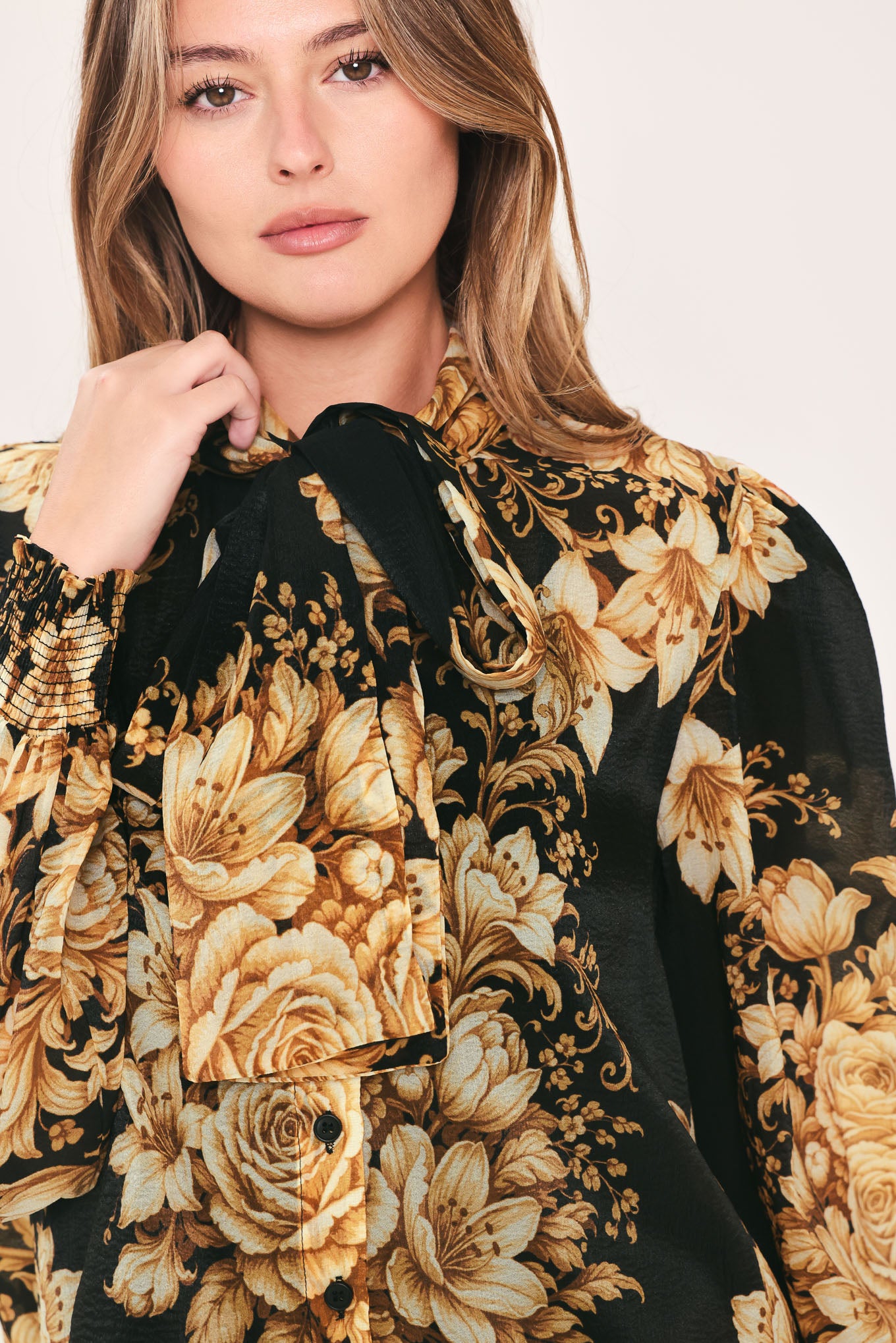 A woman wearing a black long-sleeve blouse with a large beige and gold floral print, featuring a high neck and bow detail | Flying Tomato