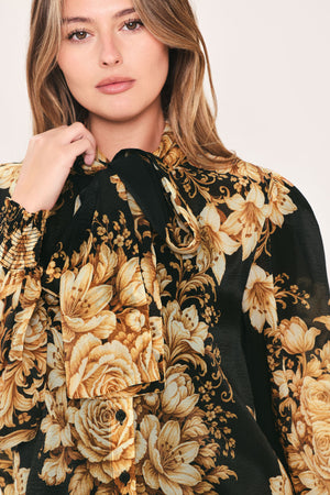 A woman wearing a black long-sleeve blouse with a large beige and gold floral print, featuring a high neck and bow detail | Flying Tomato