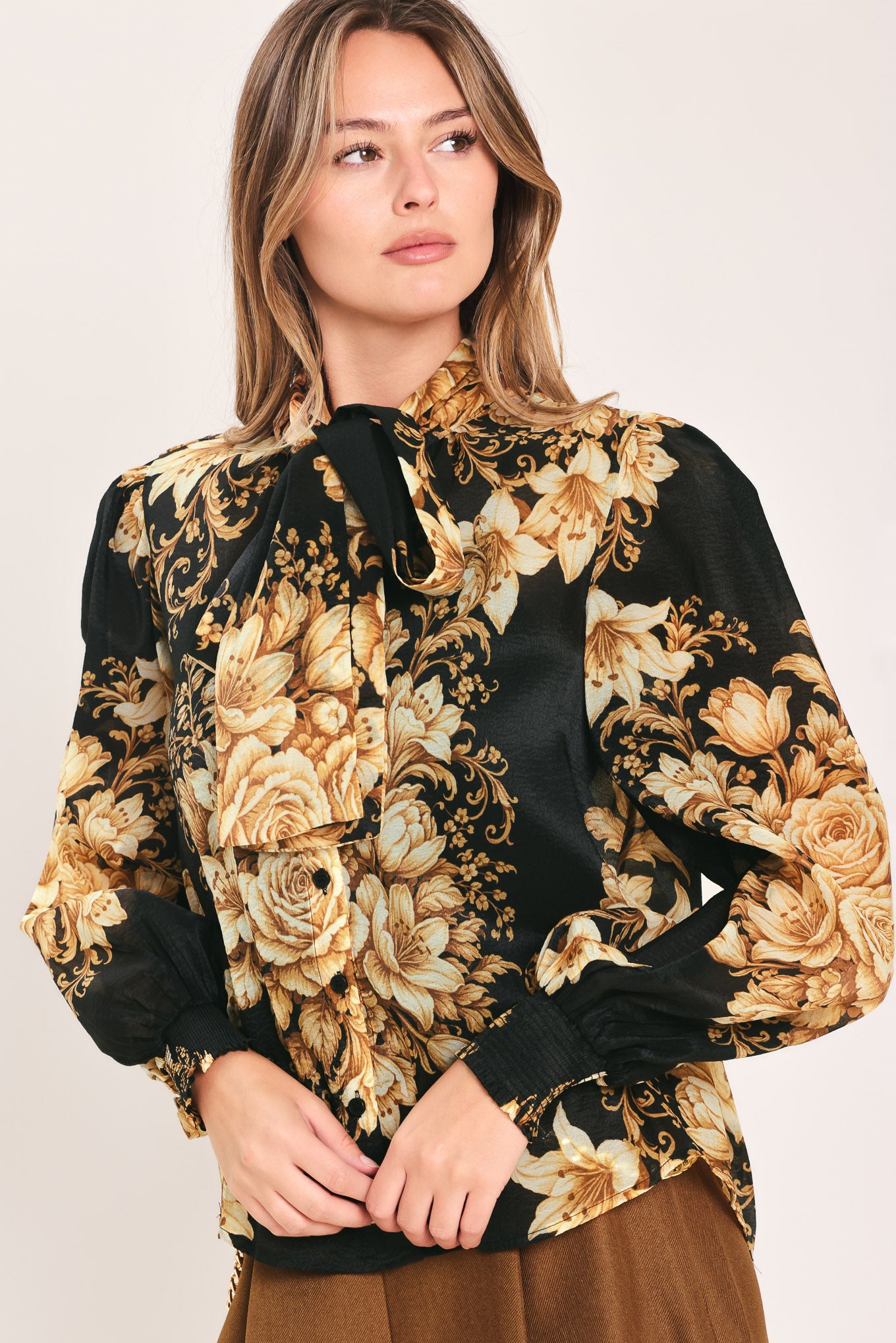 A woman wearing a black long-sleeve blouse with a large beige and gold floral print, featuring a high neck and bow detail | Flying Tomato
