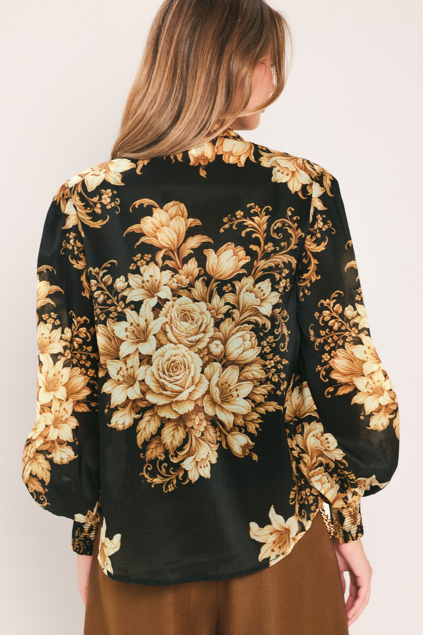 A woman wearing a black long-sleeve blouse with a large beige and gold floral print, featuring a high neck and bow detail | Flying Tomato