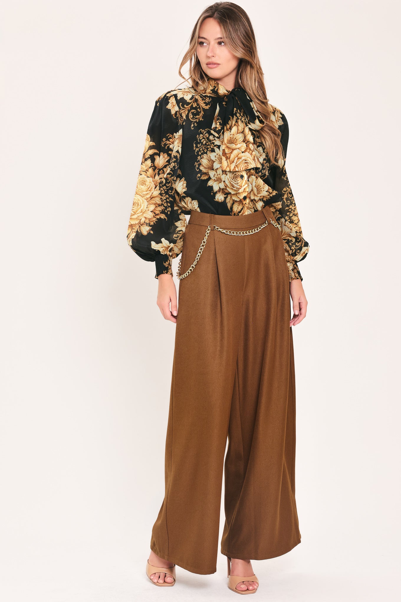 A woman wearing a black long-sleeve blouse with a large beige and gold floral print, featuring a high neck and bow detail | Flying Tomato