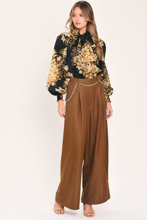 A woman wearing a black long-sleeve blouse with a large beige and gold floral print, featuring a high neck and bow detail | Flying Tomato