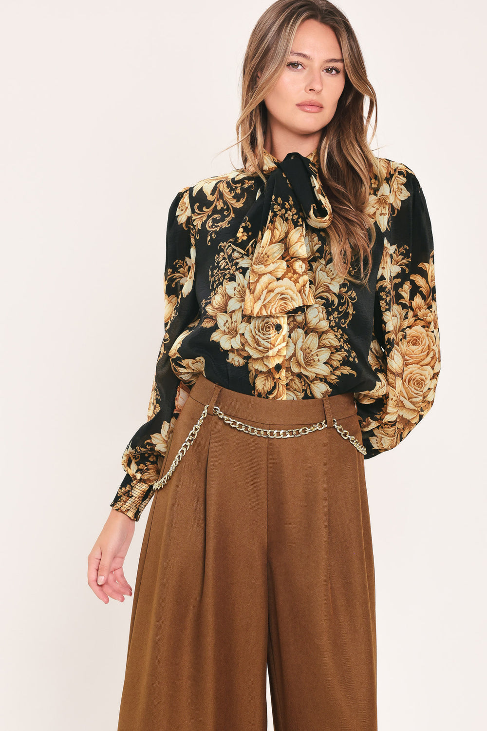 A woman wearing a black long-sleeve blouse with a large beige and gold floral print, featuring a high neck and bow detail | Flying Tomato