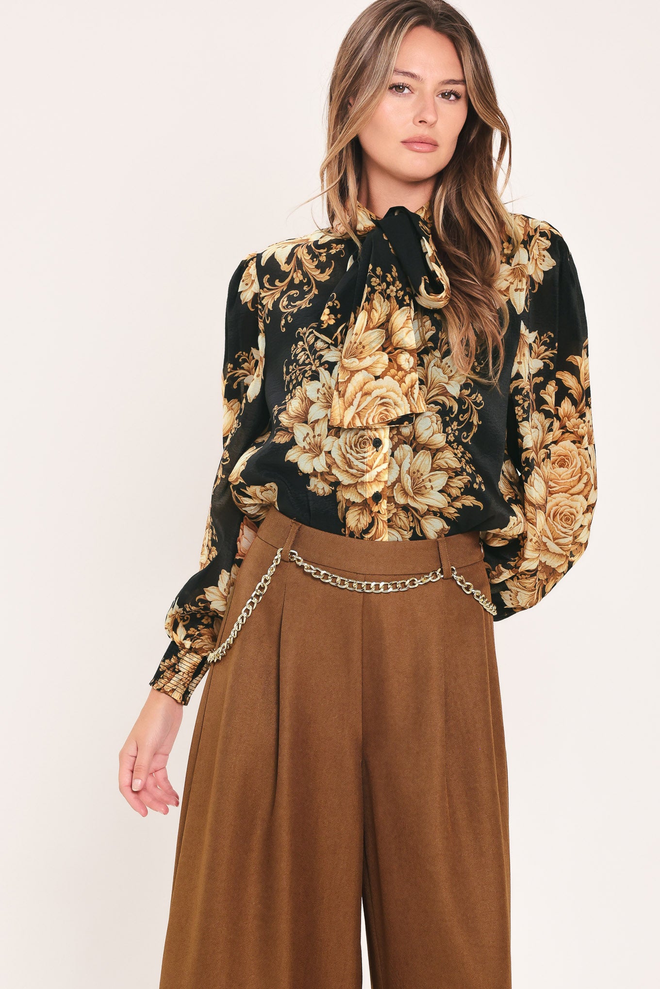 A woman wearing a black long-sleeve blouse with a large beige and gold floral print, featuring a high neck and bow detail | Flying Tomato
