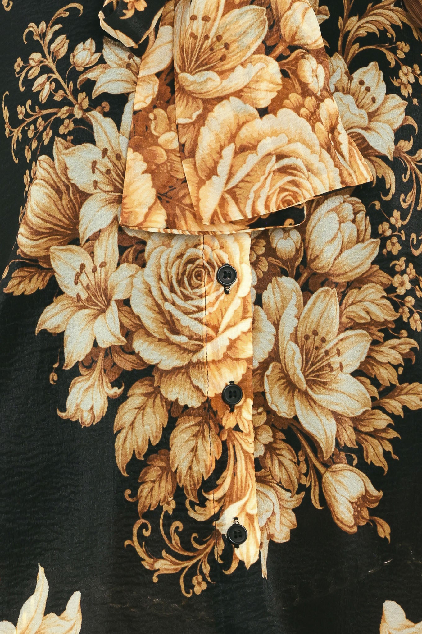 A woman wearing a black long-sleeve blouse with a large beige and gold floral print, featuring a high neck and bow detail | Flying Tomato