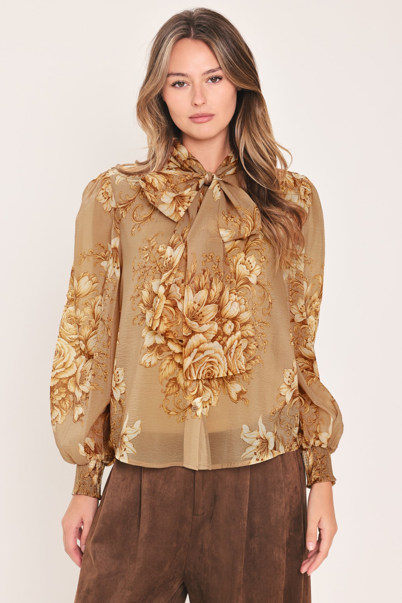 A woman wearing a sheer beige blouse with a large gold floral print and a bow tie neckline | Flying Tomato