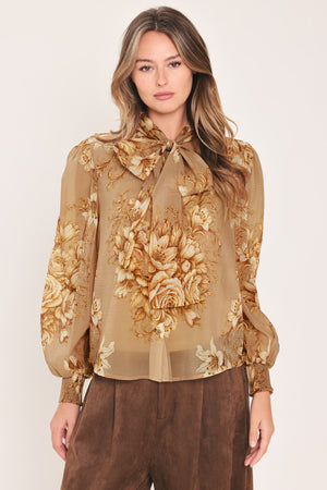 A woman wearing a sheer beige blouse with a large gold floral print and a bow tie neckline | Flying Tomato