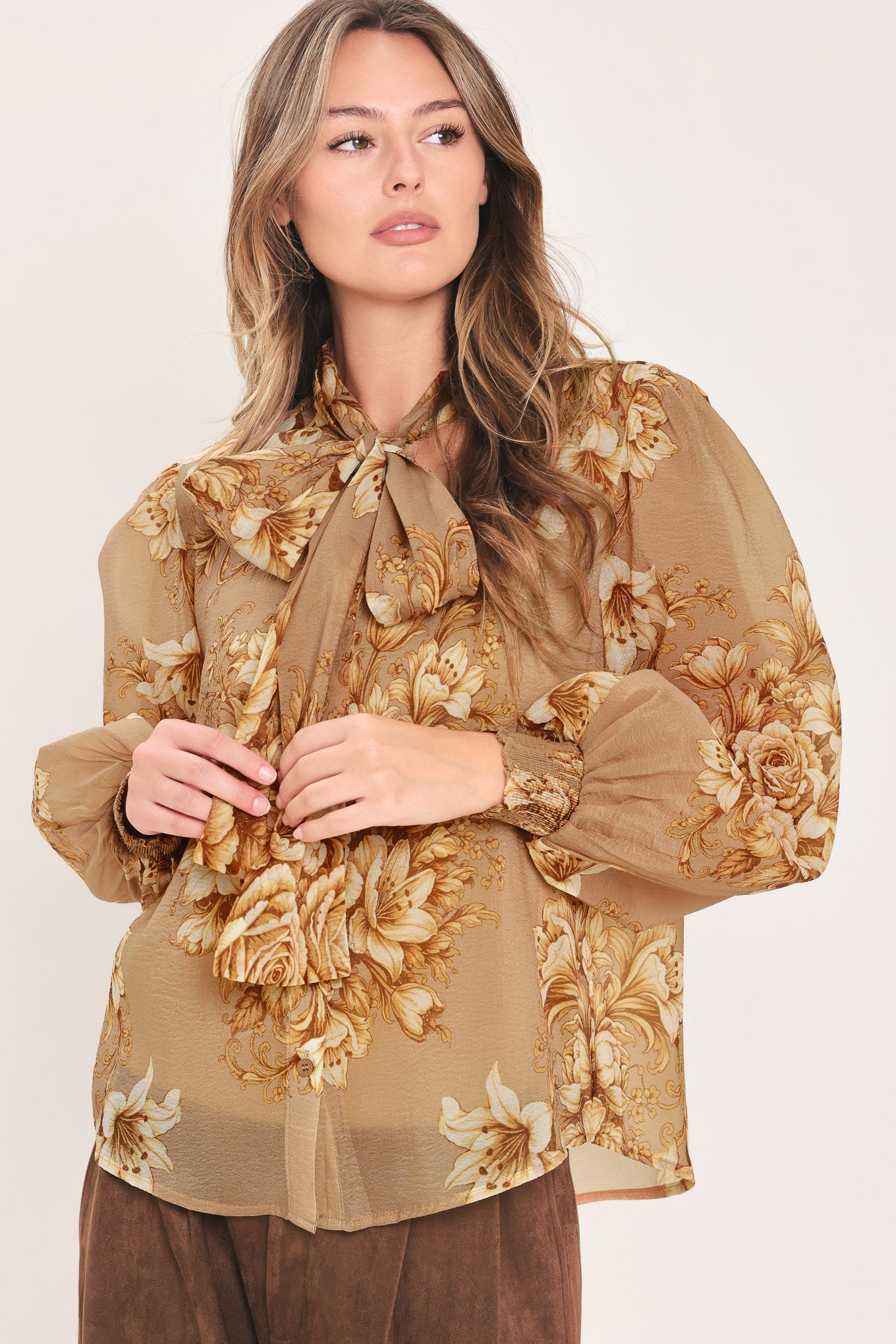 A woman wearing a sheer beige blouse with a large gold floral print and a bow tie neckline | Flying Tomato