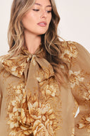 A woman wearing a sheer beige blouse with a large gold floral print and a bow tie neckline | Flying Tomato