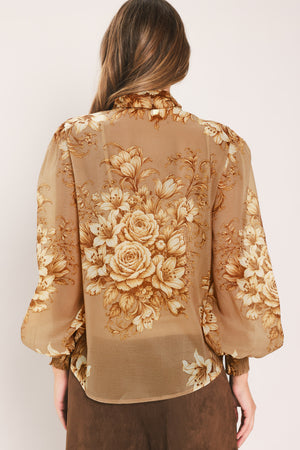 A woman wearing a sheer beige blouse with a large gold floral print and a bow tie neckline | Flying Tomato